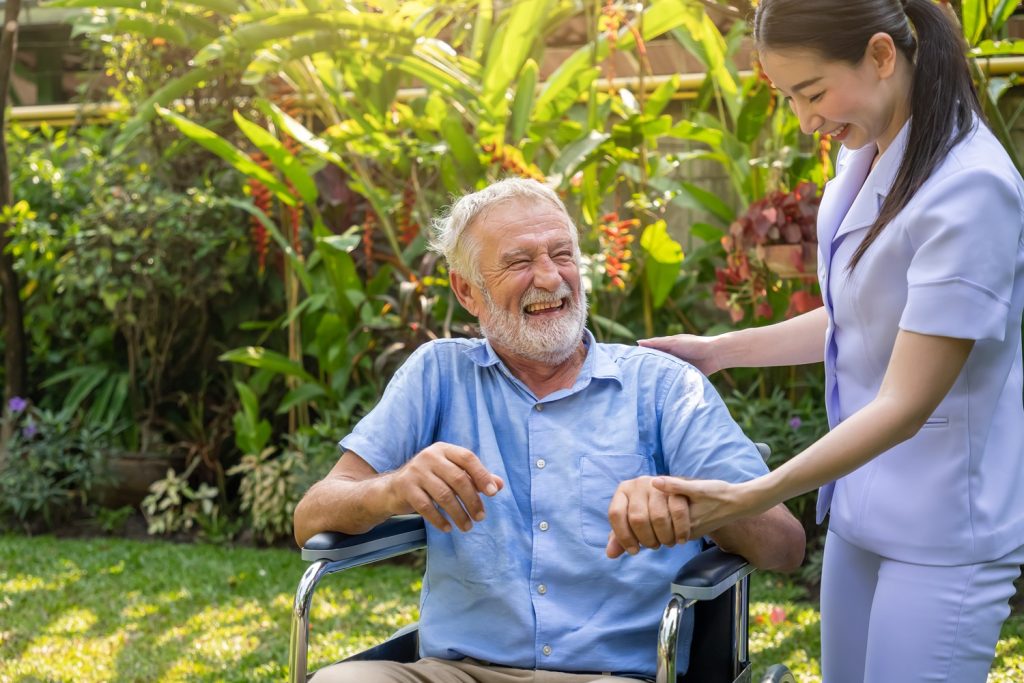 Happy nurse holding laughing elderly man hand on wheelchair in g