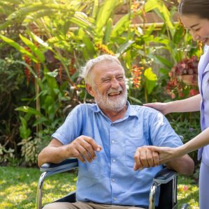 Happy nurse holding laughing elderly man hand on wheelchair in g